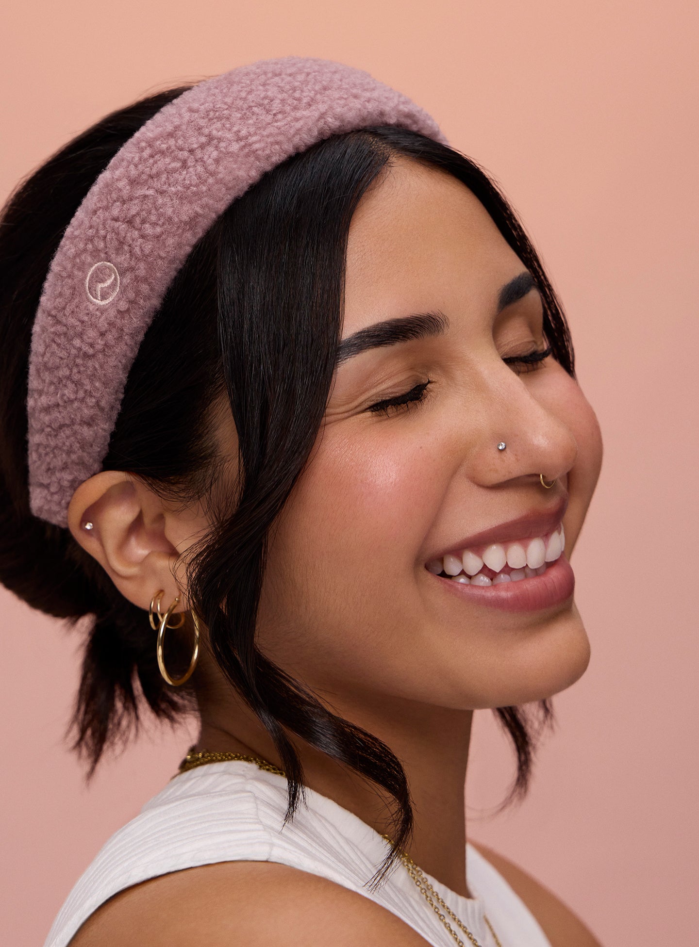 A model with black curled hair smiling and wearing Rare Beauty's Find Comfort Headband against a pink background.