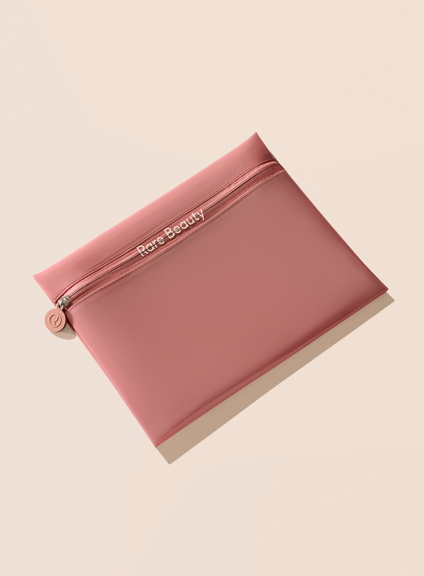 Rare Beauty's Find Comfort Tinted Clutch, a slim mauve pouch with the Rare Beauty brand name displayed on it, laying against a beige background.