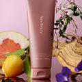 Rare Beauty's Find Comfort Gentle Exfoliating Body Wash surrounded by a lemon, vanilla beans, wood, and a grapefruit.