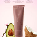 Rare Beauty's Find Comfort Gentle Exfoliating Body Wash standing on a reflective surface against a pink background next to a half an avocado, pink flowers, and a half a coconut.