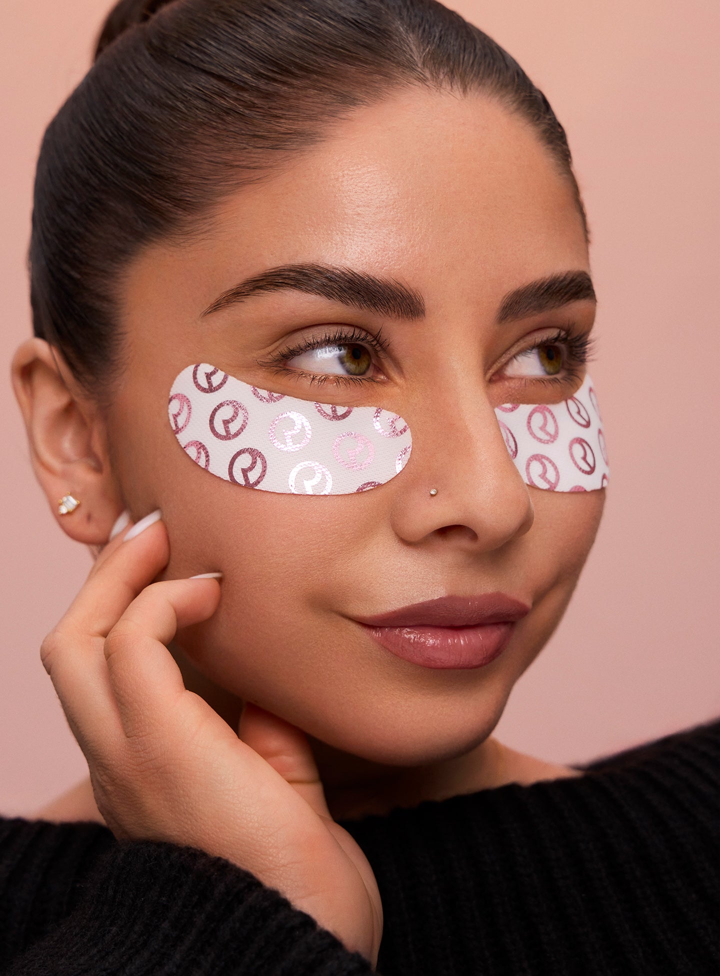 A model with a smooth bun against a pink background wearing Rare Beauty's Find Comfort Hydrating Under Eye Patches, designed to cool and smooth the under eye area for a refreshed look in minutes.