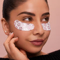 A model with a smooth bun against a pink background wearing Rare Beauty's Find Comfort Hydrating Under Eye Patches, designed to cool and smooth the under eye area for a refreshed look in minutes.