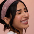 A model with black curled hair smiling and wearing Rare Beauty's Find Comfort Headband against a pink background.